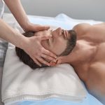 Massage therapist works with head of male lient lying on couch in contemporary clinic